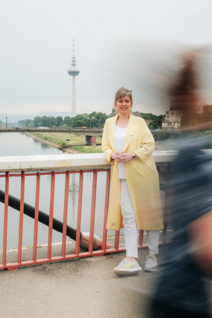 Jessica Liebe lehnt entspannt an einem Brückengeländer, während im Vordergrund eine Frau in roter Kleidung vorbeiläuft – Symbol für mentale Bewegung, Wandel und gesunde Dynamik im Berufsalltag.